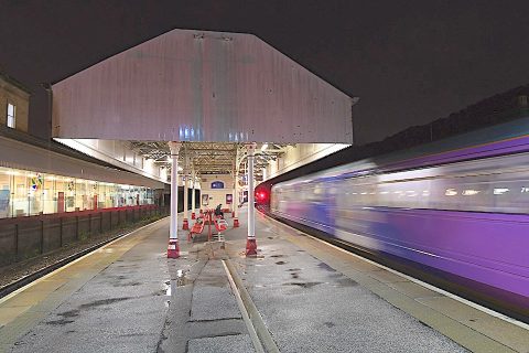 A night time view looking down a station platform at Halifax with a red signal in the distance and a fast train blurring by