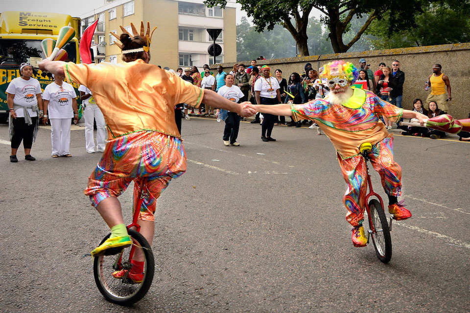 Two unicyclists in multicoloured costumes circle each other while shaking hands