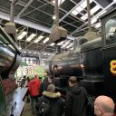 A historic railway locomotive roundhouse, with a crowd of people walking between two steam locomotives and approaching an electric shunting locomotive rebuilt from a class 08 diesel