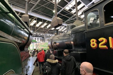 A historic railway locomotive roundhouse, with a crowd of people walking between two steam locomotives and approaching an electric shunting locomotive rebuilt from a class 08 diesel