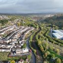 An aerial view of Ebbw Vale with rows of houses running north south up the picture on the left, and a road and railway winding up the valley divides the picture. There is a modern large Industrail building on the right. The sky is overcast