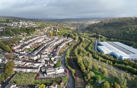 An aerial view of Ebbw Vale with rows of houses running north south up the picture on the left, and a road and railway winding up the valley divides the picture. There is a modern large Industrail building on the right. The sky is overcast