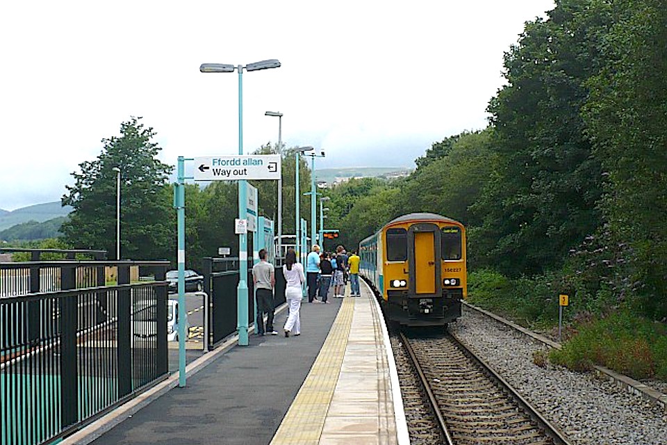 Diesel train arriving at Rogerstone station in rural Wales. A few people stand on the single platform. There are trees all around