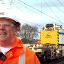 Candid portrait of a smiling Jemery Spence of Network Rail in orange coat in front of a yellow Kirow track crane